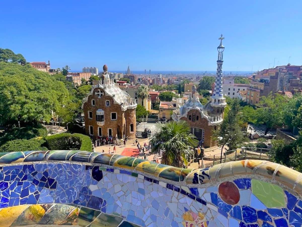 View from Park Güell in Barcelona, showing Gaudí’s whimsical mosaic terrace in the foreground, two colorful gingerbread-style gatehouses below, and the Sagrada Familia and Mediterranean Sea in the distant skyline.