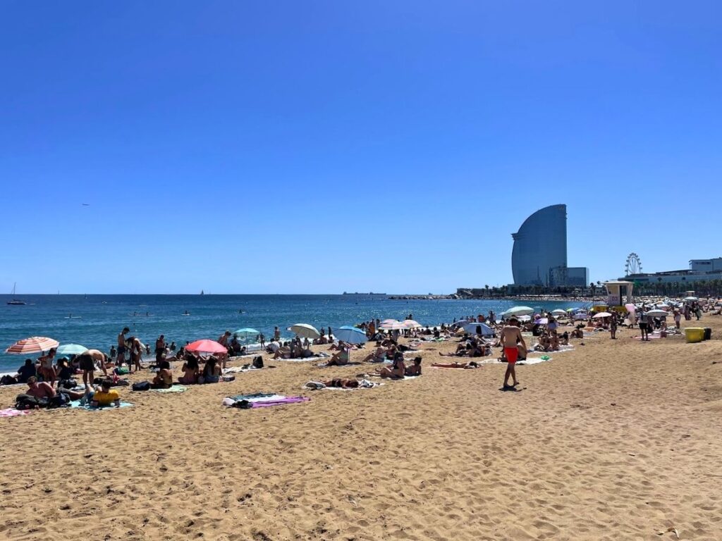 Crowded Barceloneta Beach with colorful umbrellas, sunbathers, and the W Barcelona hotel in the distance