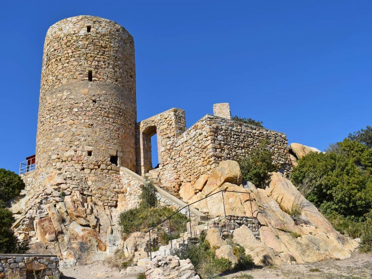 Ruins of Castell de Burriac near Cabrera de Mar, Catalonia, featuring a round medieval stone watchtower, crumbling fortress walls, and rocky steps under a clear blue sky