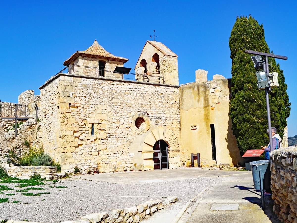 Historic stone chapel and bell tower inside Castell de la Santa Creu de Calafell in Spain, featuring an arched entrance, medieval walls, and a tall cypress tree