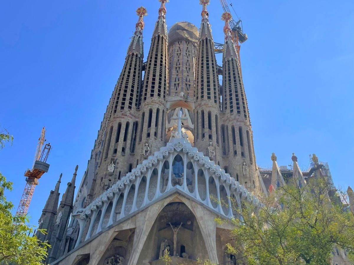 Close-up view of La Sagrada Familia's Passion Facade in Barcelona, featuring towering spires, intricate stone carvings, and a crucifixion sculpture against a clear blue sky