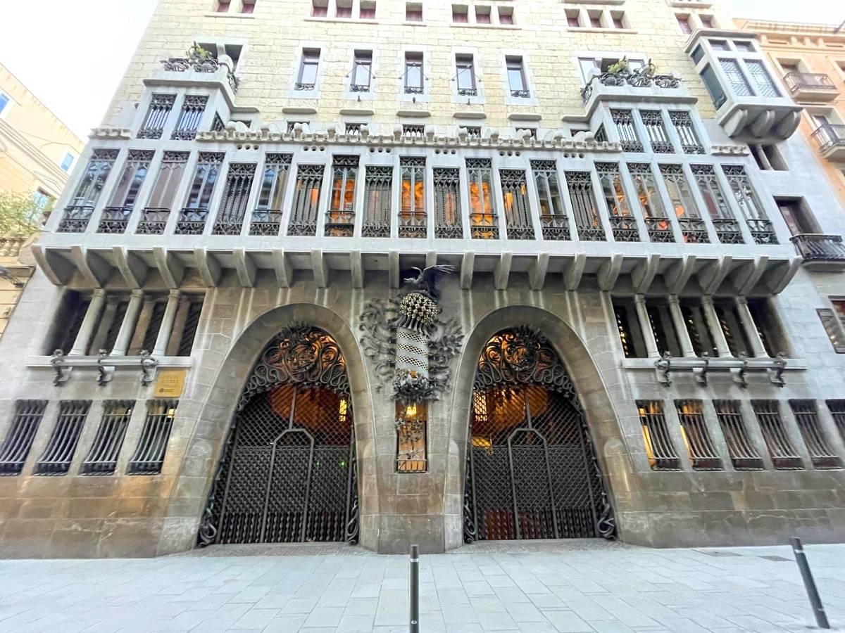 
Façade of Palau Güell in Barcelona, designed by Antoni Gaudí, featuring grand parabolic arches, intricate wrought iron detailing, and vertical window grilles across the upper floors, all in a stately stone structure.