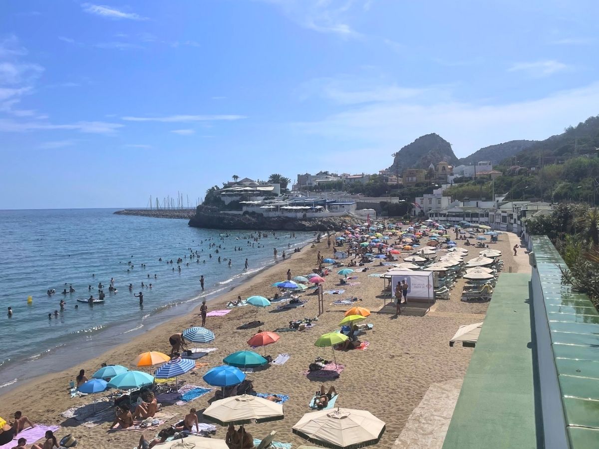 Crowded sandy beach in Garraf, Spain with colorful umbrellas, sunbathers, and swimmers in the calm blue sea under a clear sky