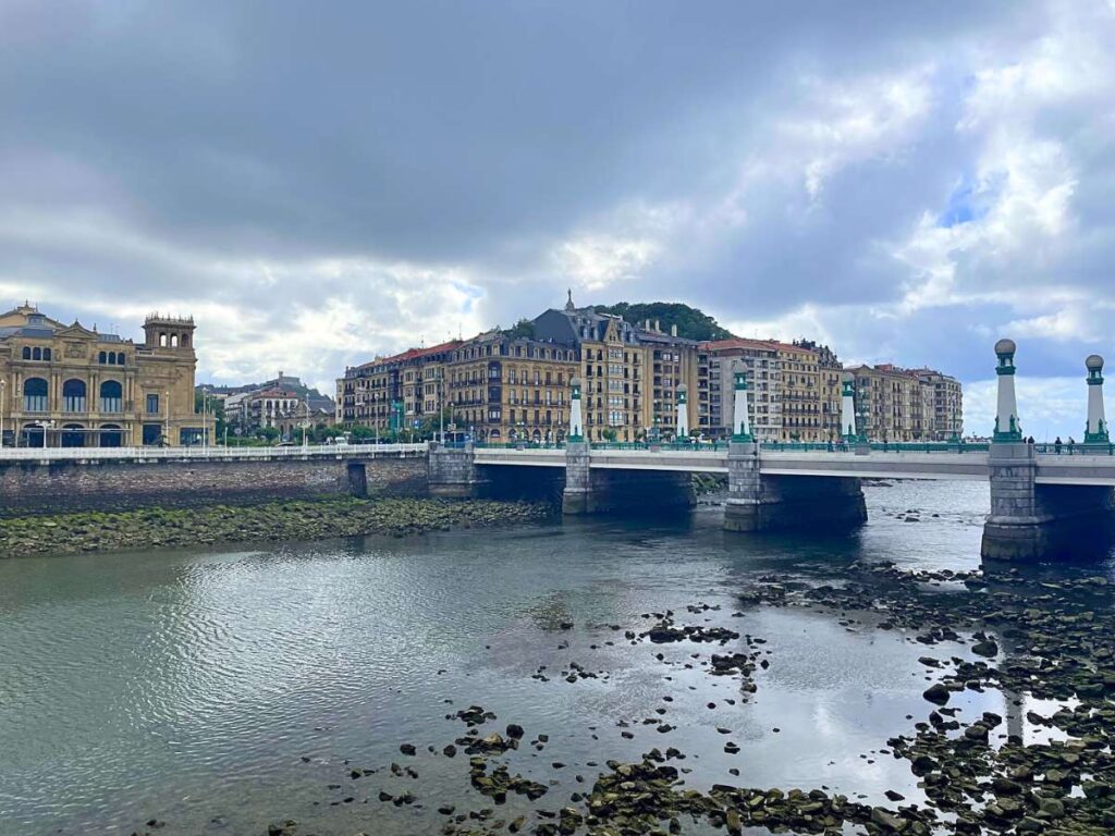 View of the Kursaal Bridge crossing the Urumea River in San Sebastian, Spain, with historic waterfront buildings and cloudy skies in the background