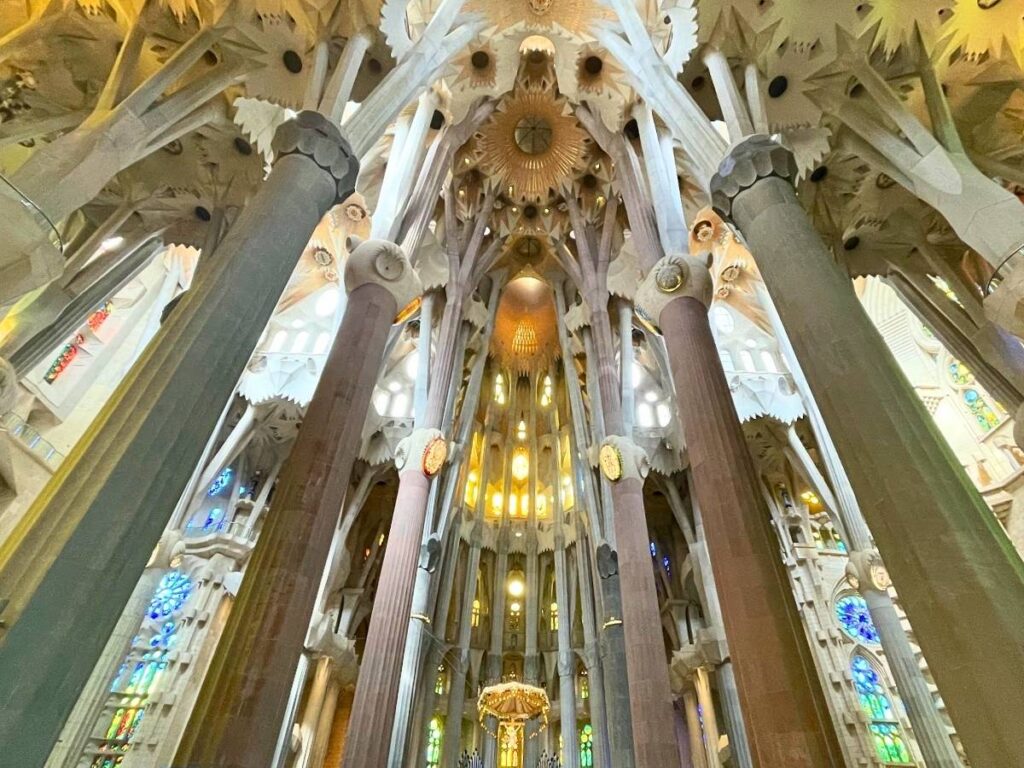 Interior of La Sagrada Familia in Barcelona, featuring towering tree-like columns, intricate vaulted ceilings, colorful stained glass windows, and the illuminated altar