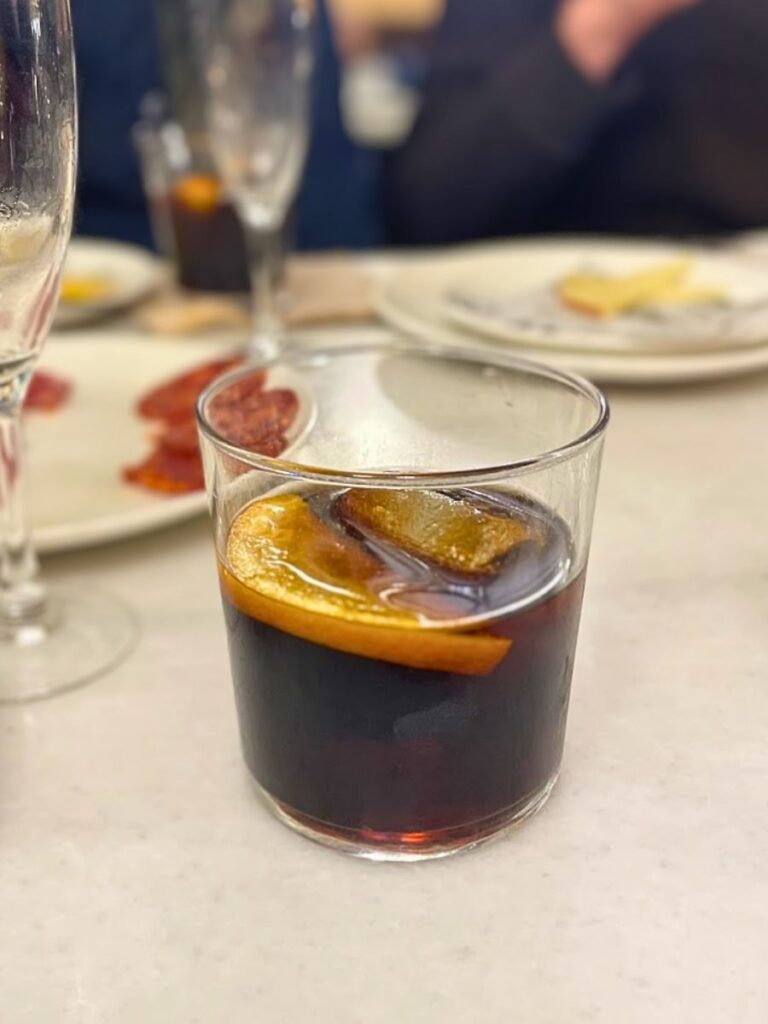 A glass of dark vermouth with an orange slice sits on a table surrounded by tapas dishes