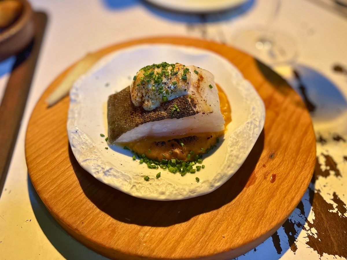 A thick fillet of fish topped with a dollop of sauce and chives, served over a golden purée on a ceramic plate placed on a wooden board

