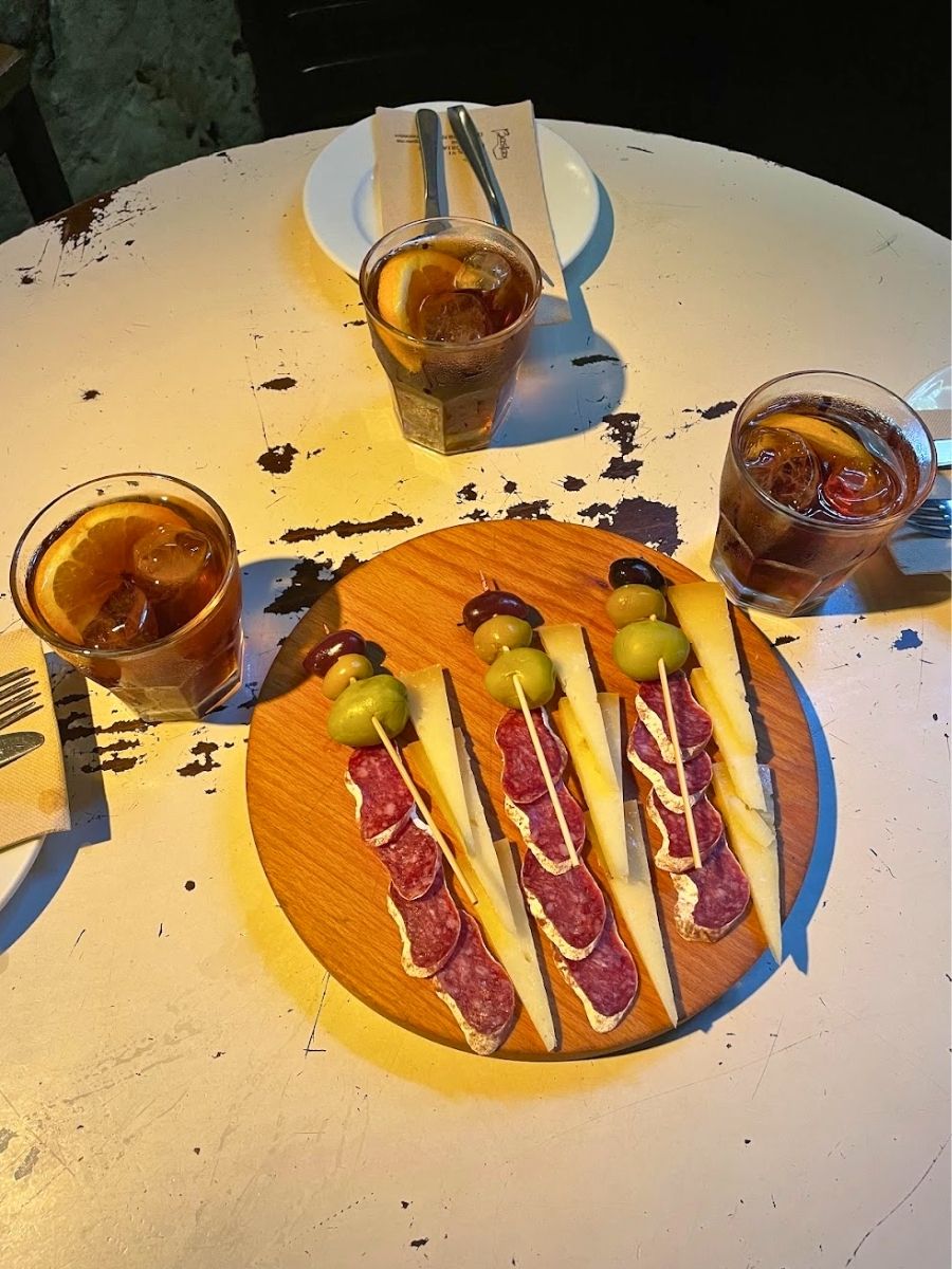 A rustic round wooden board with cheese slices, salami, olives, and drizzle on breadsticks, accompanied by three glasses of vermouth with orange slices on a distressed white table