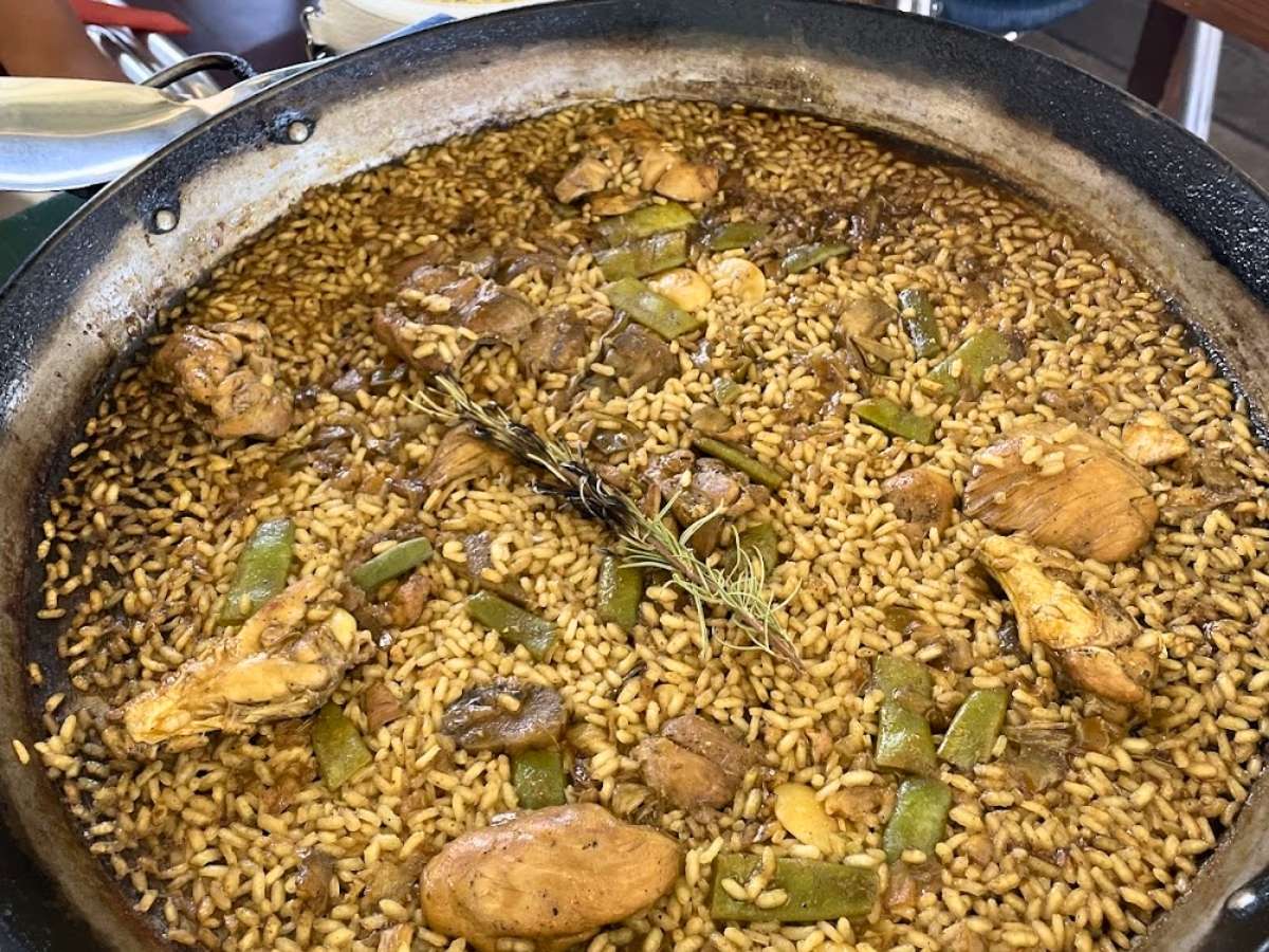 Large pan of paella with golden rice pieces of chicken green beans and mushrooms topped with a sprig of rosemary cooking in a wide shallow pan