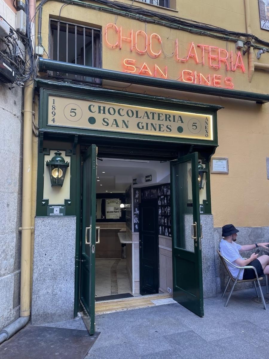 Entrance of the iconic Chocolatería San Ginés with neon signage and a man seated outside under the open green doors