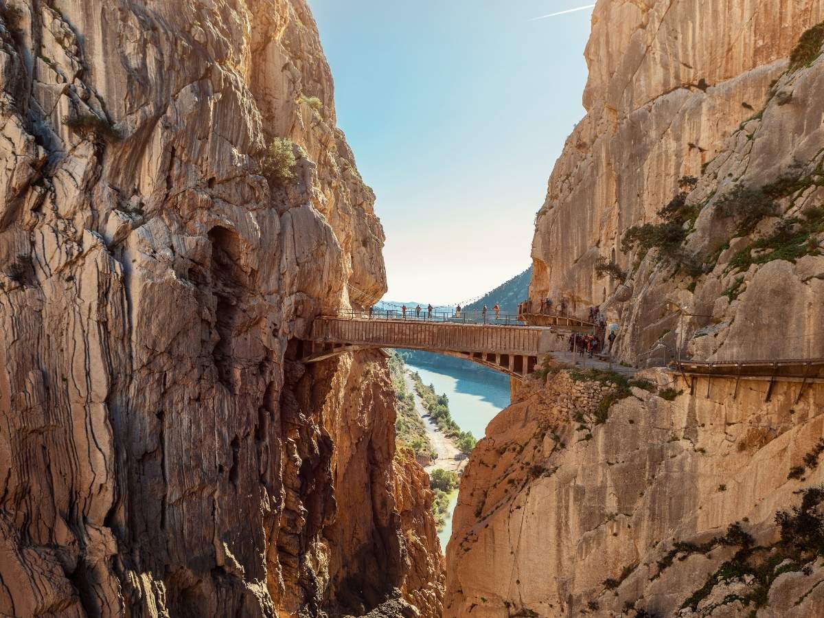  Hikers walking along El Caminito del Rey, a narrow pathway clinging to steep rock cliffs above a deep gorge in southern Spain, with a river winding below and sunlight filtering between the canyon walls.