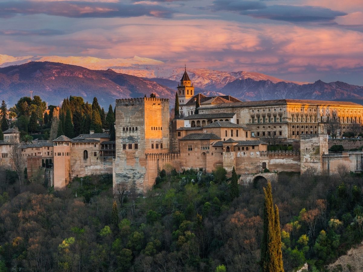 The Alhambra palace and fortress complex in Granada, Spain, bathed in golden evening light, with the snow-capped Sierra Nevada mountains in the background and lush green forest in the foreground.