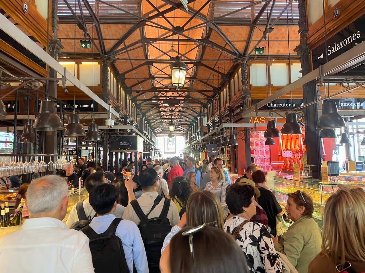 Crowds fill the vibrant San Miguel Market in Madrid, surrounded by gourmet food stalls offering jamón, wine, and tapas under an ornate iron-and-glass roof