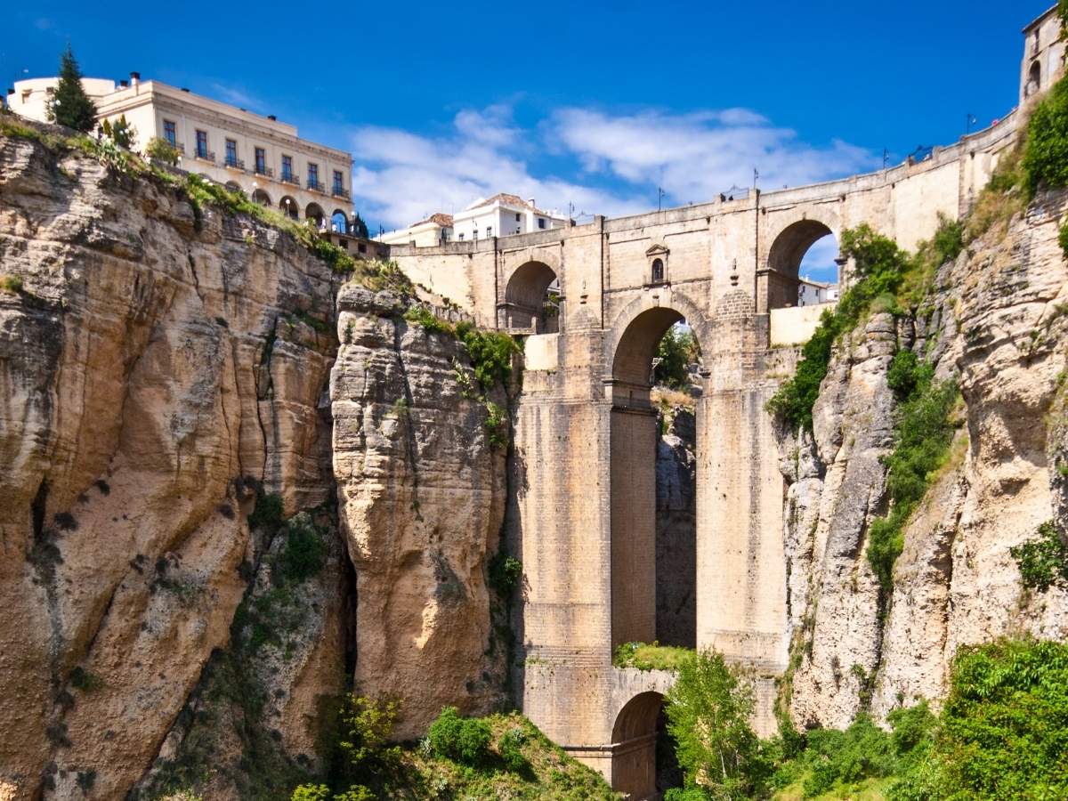 The Puente Nuevo bridge in Ronda, Spain, dramatically spanning a deep gorge with rugged cliffs, connecting the historic city on either side under a bright blue sky.