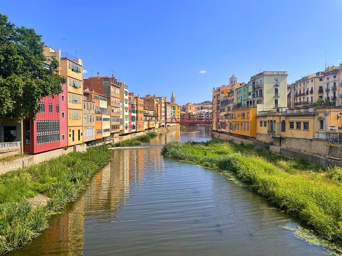 Riverside view of colorful buildings along the Onyar River in Girona Spain with a red iron bridge in the distance