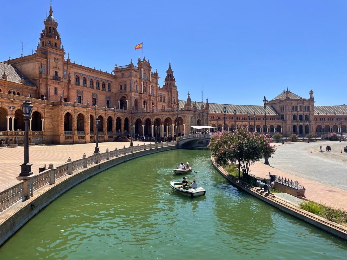 People rowing small boats along the canal in front of the grand, arched buildings of Plaza de España on a sunny day in Seville