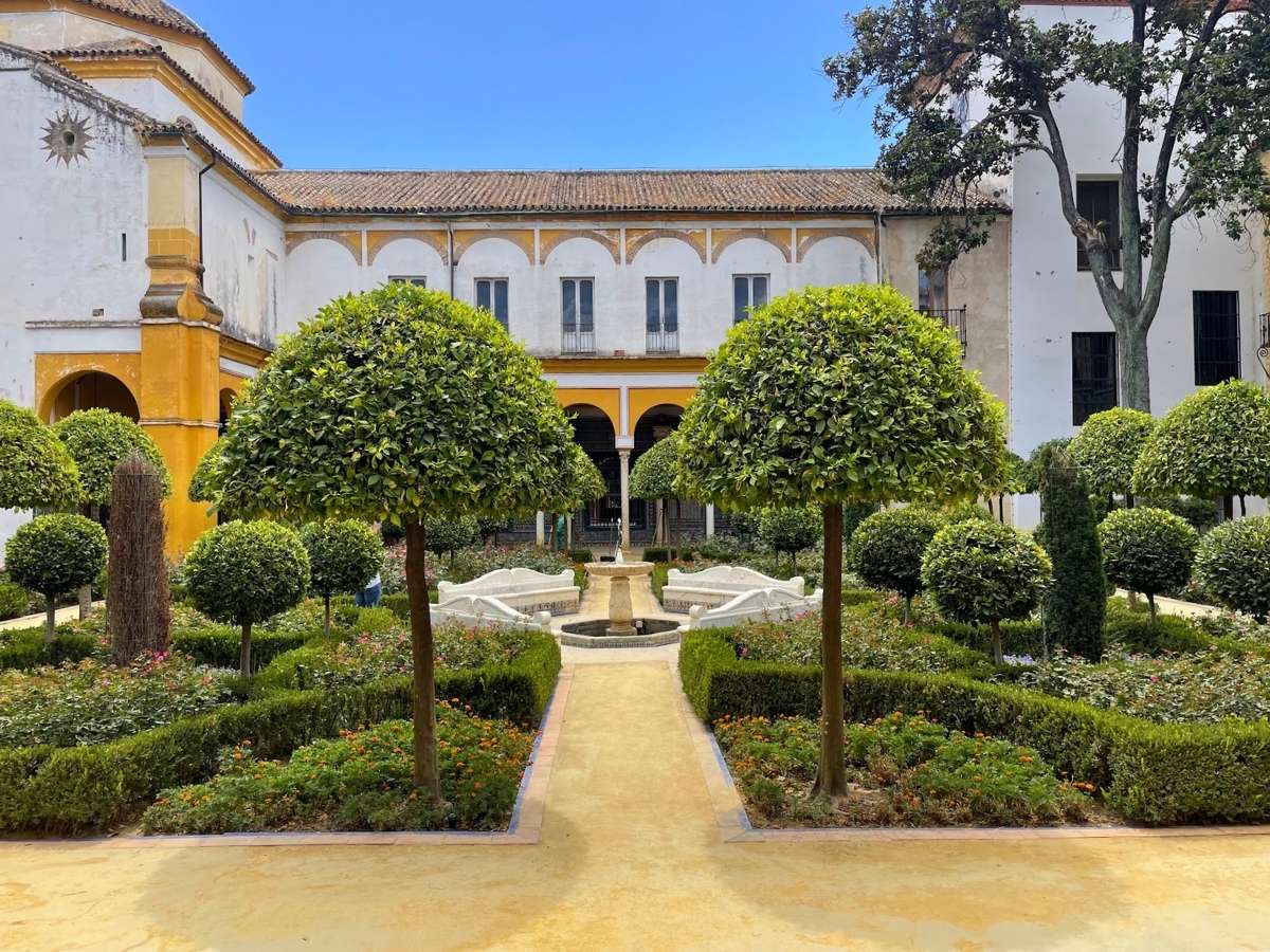 A manicured courtyard garden at Casa de Pilatos in Seville with neatly trimmed trees, flowerbeds, and a central stone fountain framed by a white and yellow building