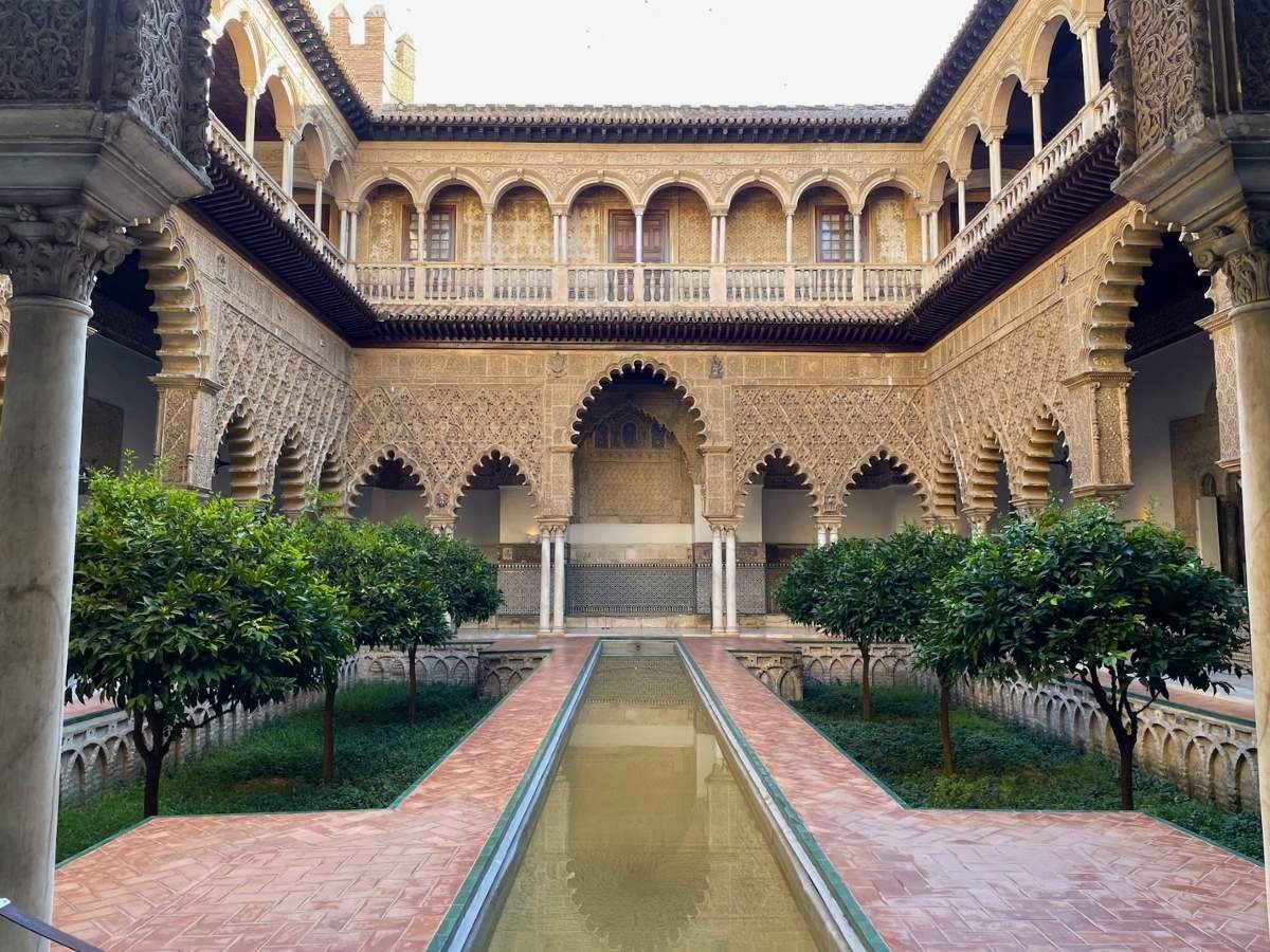 An ornate courtyard in the Real Alcázar of Seville featuring Islamic-style arches, intricate wall carvings, and a long rectangular reflecting pool