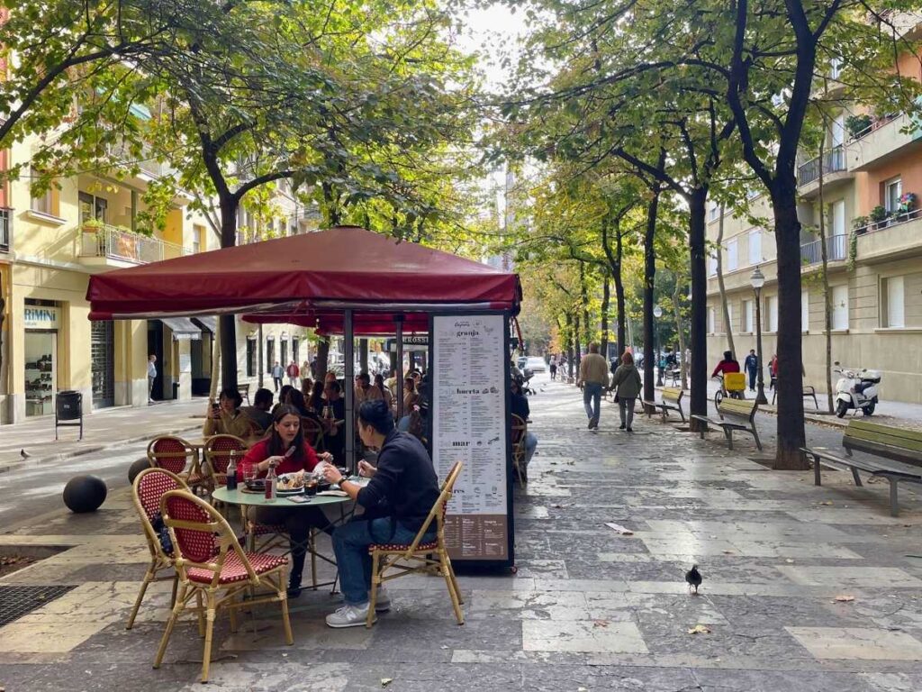 Outdoor café terrace with red chairs and a burgundy canopy on a tree-lined pedestrian street in Barcelona, with people dining and walking nearby