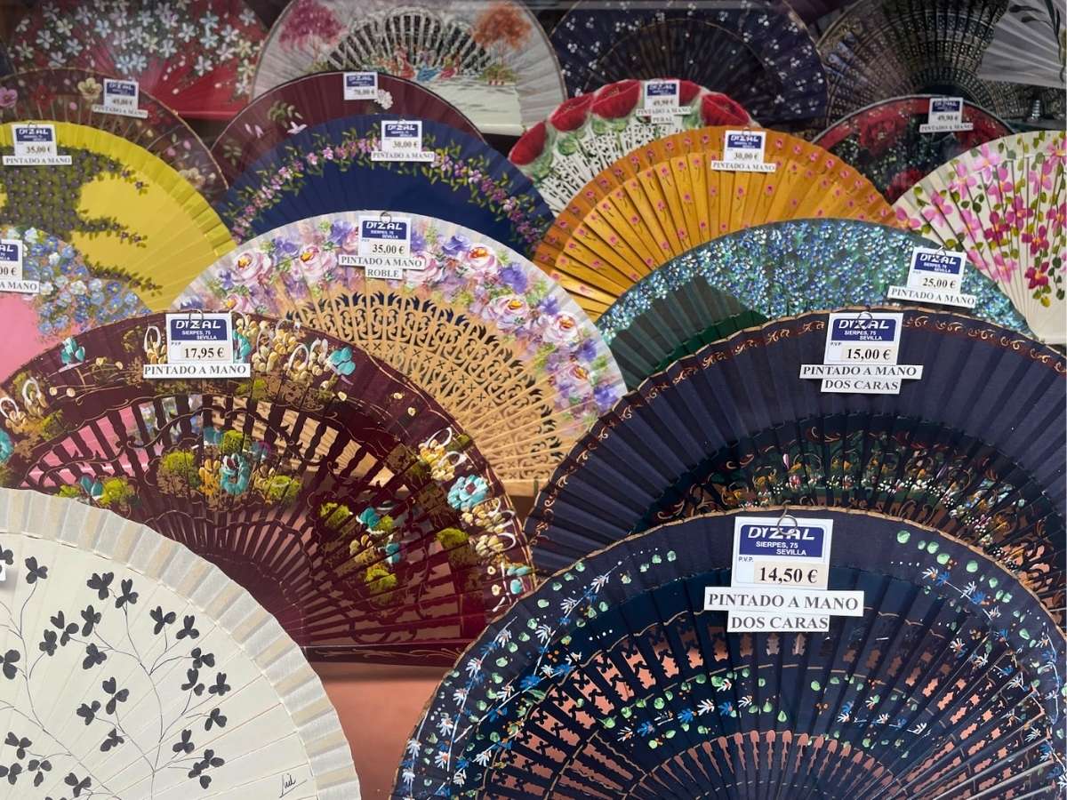 Hand-painted folding fans in vibrant patterns and colors arranged in a shop window display with visible price tags