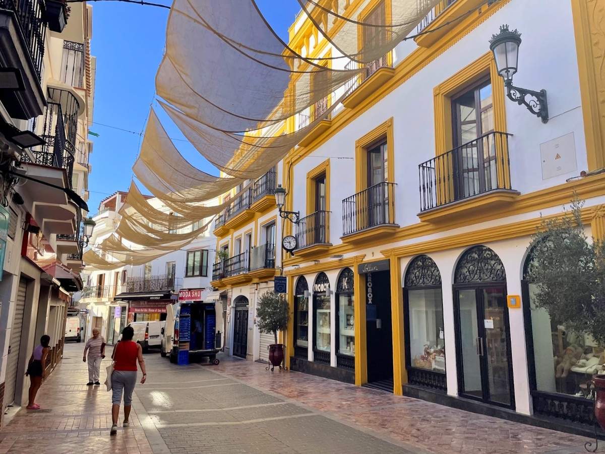 Narrow pedestrian shopping street with yellow-trimmed buildings and sunshades overhead in Nerja Spain