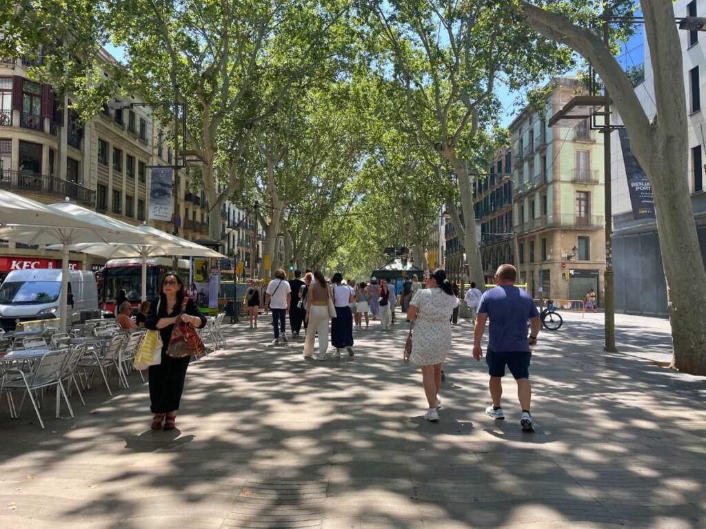 Pedestrians walking along the wide tree lined La Rambla in Barcelona with outdoor café seating and historic buildings on both sides