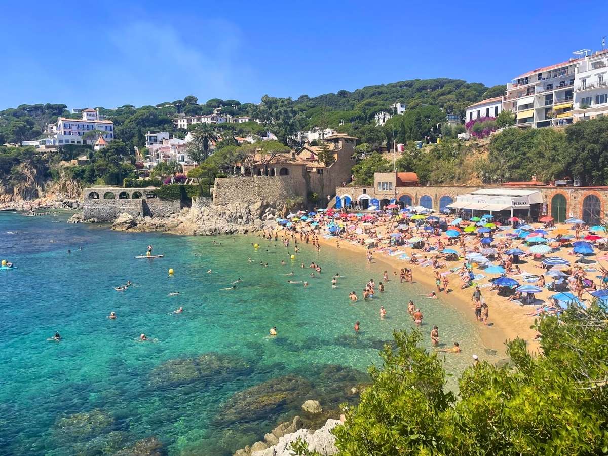 Beachgoers swimming and relaxing on the sandy beach in Calella de Palafrugell beside colorful waterfront buildings and clear turquoise water