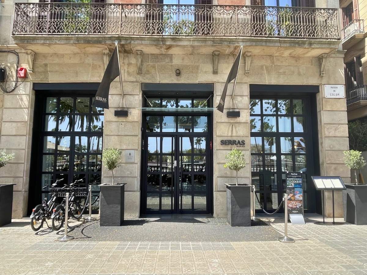 Entrance of Serras Barcelona hotel with black-framed glass doors and facade facing Passeig de Colom