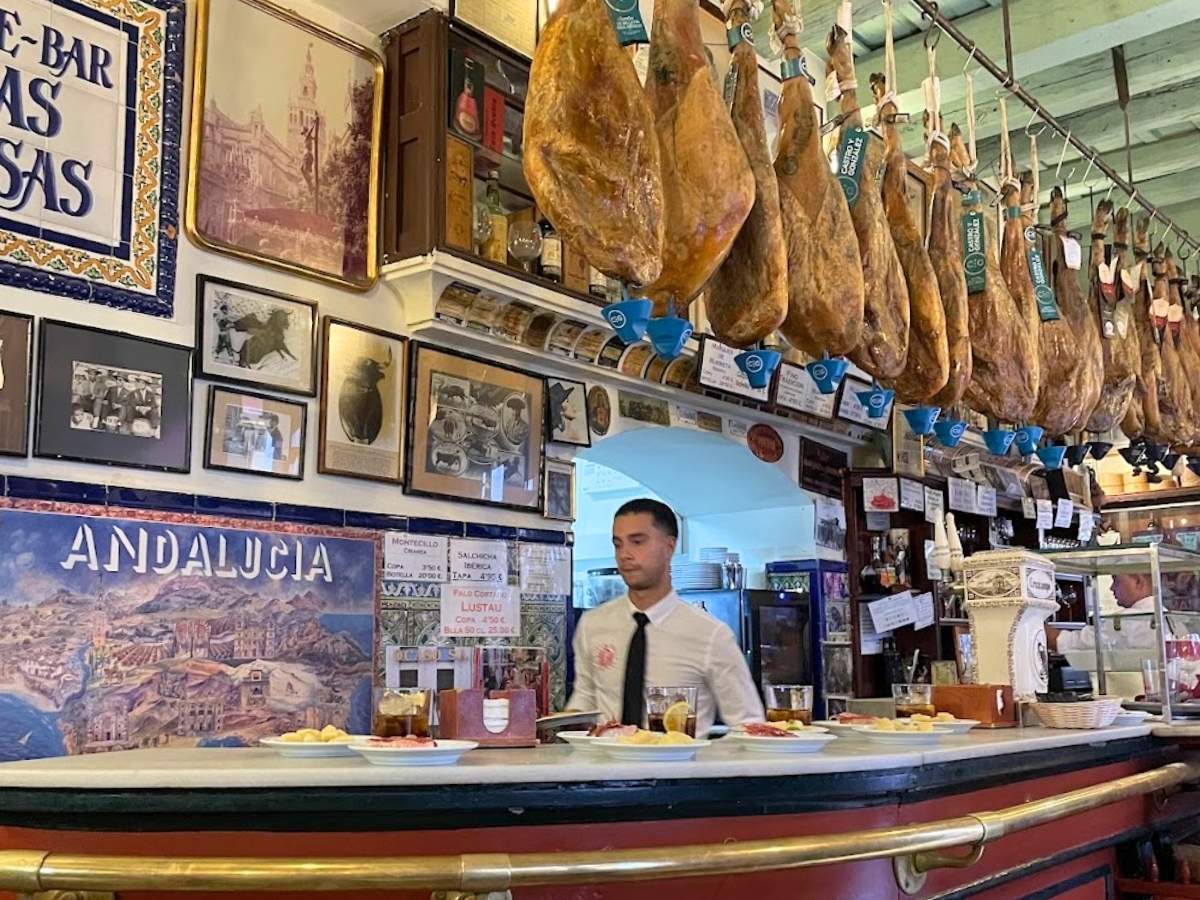Bartender standing behind the counter of a traditional Seville tapas bar with hanging jamón ibérico and Spanish wall decorations