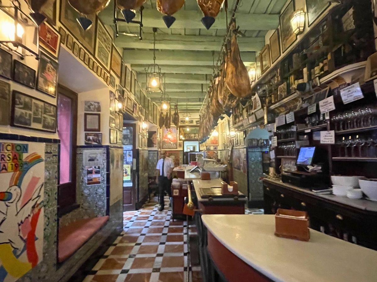 Interior of a traditional Seville tapas bar with hanging cured hams, tiled floors, and vintage photographs