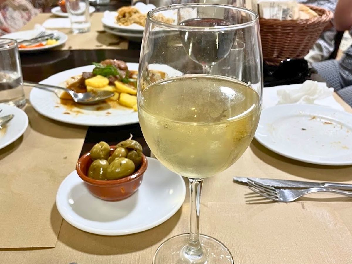 Glass of white wine with a small bowl of green olives and traditional Spanish tapas in the background