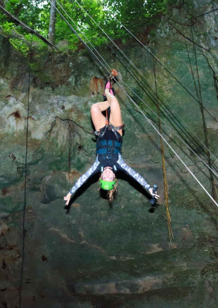 Me wearing a green helmet and safety harness hanging upside down on a zipline above a rocky cenote in Mexico